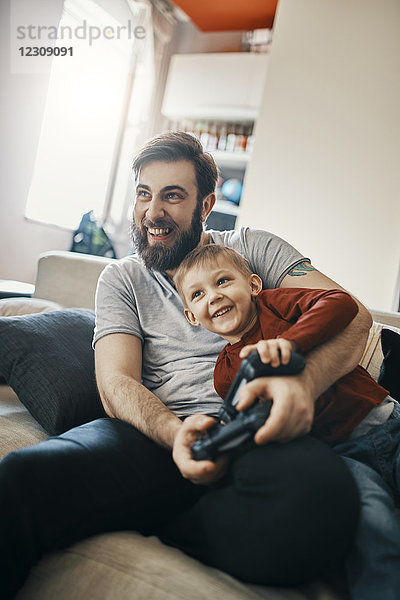 Lachender Vater und kleiner Sohn sitzen zusammen auf der Couch und spielen ein Computerspiel.