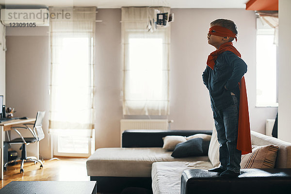 Kleiner Junge verkleidet als Superheld auf dem Wohnzimmertisch stehend.