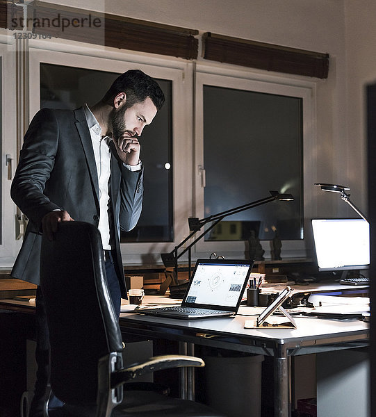 Geschäftsmann steht nachts im Büro und schaut auf den Laptop.