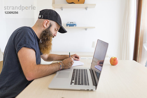 Ein bärtiger junger Mann sitzt am Schreibtisch mit einem Laptop  der etwas aufschreibt.