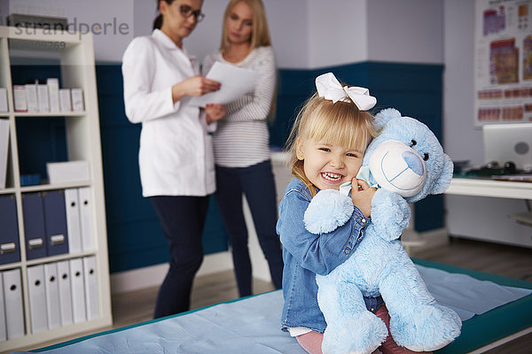 Mädchen umarmender Teddy in der Arztpraxis mit Arzt und Mutter im Hintergrund