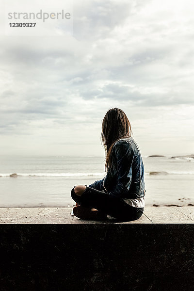 Junge Frau sitzt auf einer Mauer und schaut aufs Meer.