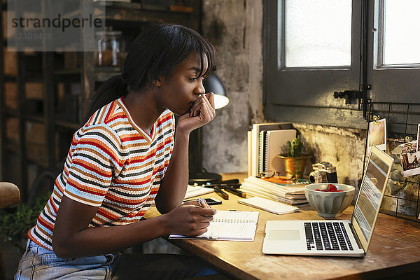 Nachdenkliche junge Frau sitzt am Schreibtisch in einem Loft und schaut auf den Laptop.