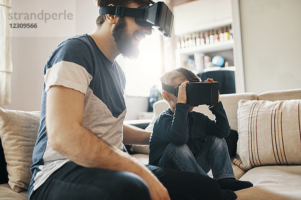 Vater und kleiner Sohn sitzen zu Hause auf der Couch und tragen eine Virtual-Reality-Brille.