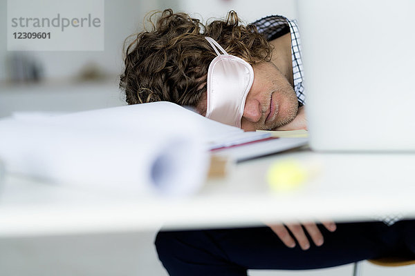 Überarbeiteter Geschäftsmann  der an seinem Schreibtisch schläft und eine Schlafmaske trägt.