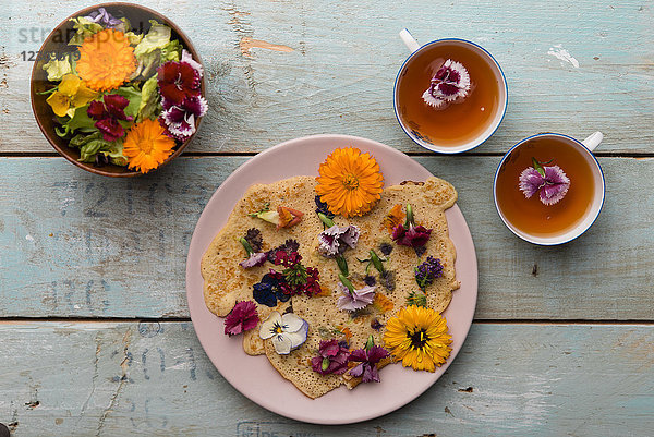 Pfannkuchen mit essbaren Blüten  Kürbisblüten  Calendula  Chamaemelum nobile  Dianthus  Taraxacum officinale  Viola  Rosmarinus officinalis auf Bambusplatte und Tassen Tee