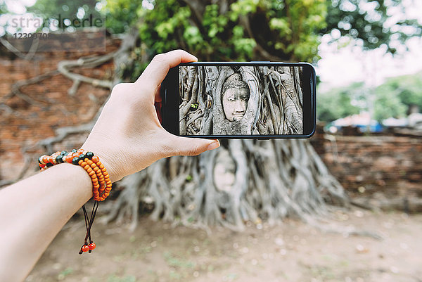 Thailand  Ayutthaya  Wat Mahathat  Frau beim Fotografieren mit dem Smartphone des Buddhakopfes in Baumwurzeln