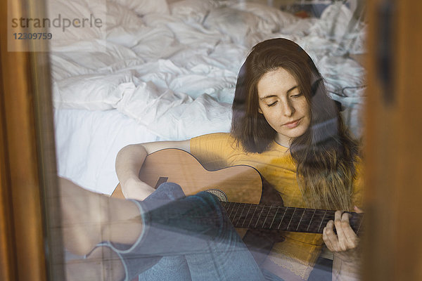 Porträt einer entspannten jungen Frau  die hinter einer Fensterscheibe sitzt und Gitarre spielt.