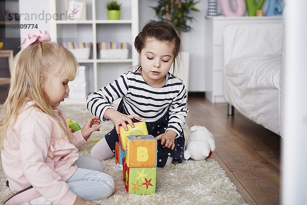 Zwei kleine Mädchen  die zu Hause mit Spielzeugblöcken auf dem Teppich spielen.