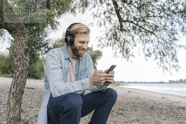 Deutschland  Düsseldorf  lächelnder Mann mit Kopfphobien über Smartphone am Strand
