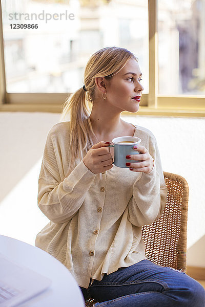 Blonde Frau mit einer Tasse Kaffee  die aus dem Fenster schaut.