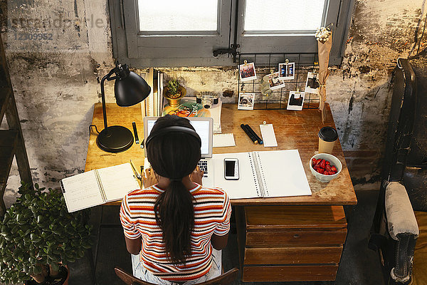 Rückansicht einer jungen Frau am Schreibtisch in einem Loft  die von oben gesehen am Laptop arbeitet.