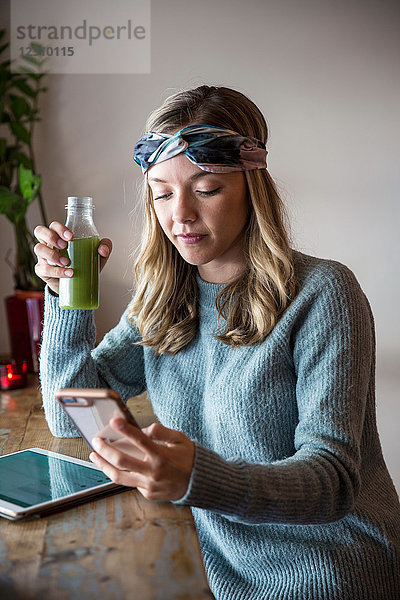 Junge Frau mit Gemüsesaft betrachtet Smartphone am Fensterplatz eines Cafés