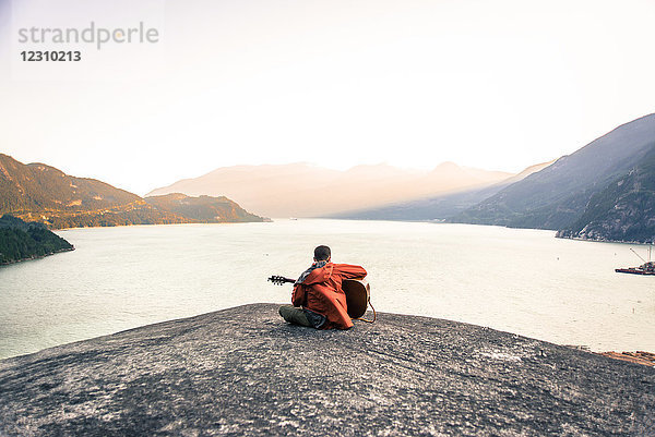 Junger Mann sitzt auf dem Malamute  spielt Gitarre  Rückansicht  Squamish  Kanada