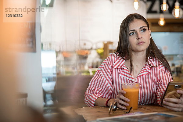 Junge Frau sitzt im Café  hält Smartphone in der Hand und trinkt Smoothie