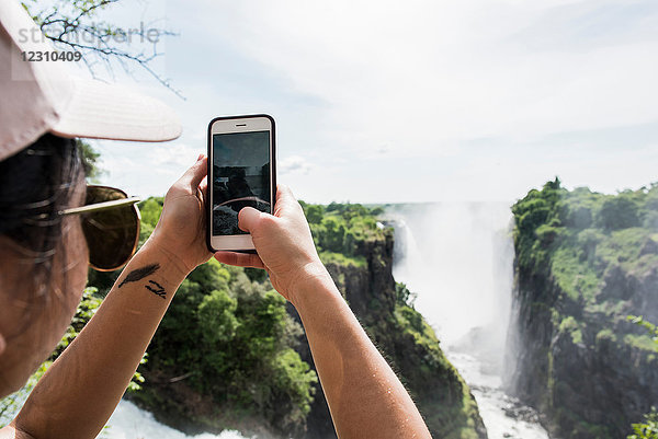 Junge Touristin beim Fotografieren der Victoria-Fälle per Smartphone  über die Schulter gesehen  Simbabwe  Afrika