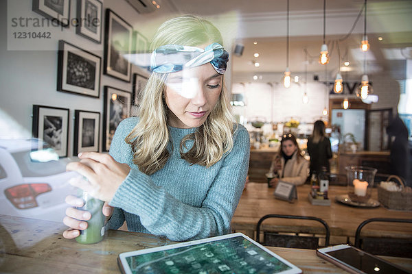 Junge Frau mit Gemüsesaft betrachtet digitales Tablett am Fensterplatz eines Cafés