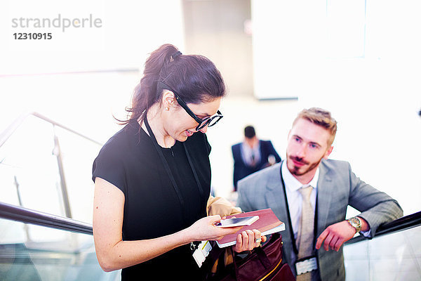 Junge Geschäftsfrau und Mann auf der Rolltreppe am Flughafen schauen auf Smartphone