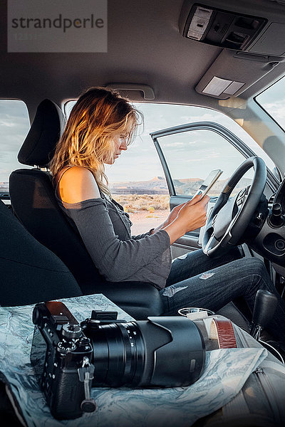 Junge Frau sitzt im Fahrzeug und schaut auf ein digitales Tablett  Spiegelreflexkamera auf dem Beifahrersitz  Mexican Hat  Utah  USA