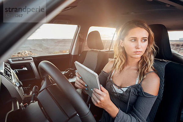 Junge Frau sitzt im Fahrzeug  hält ein digitales Tablet und schaut aus dem Fenster  Mexican Hat  Utah  USA