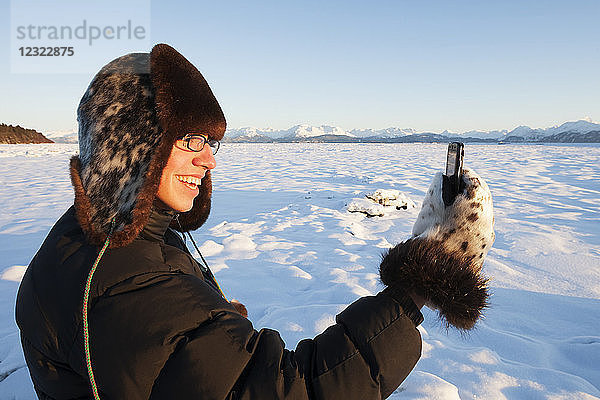 Mann mit Pelzmütze und Fäustlingen steht im Schnee und telefoniert  Süd-Zentral-Alaska; Alaska  Vereinigte Staaten von Amerika