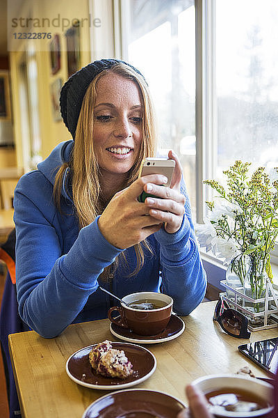 Junge Frau genießt eine Tasse Tee und Gebäck  während sie in einem Café eine SMS auf ihrem Smartphone schreibt  Süd-Zentral-Alaska; Anchorage  Alaska  Vereinigte Staaten von Amerika