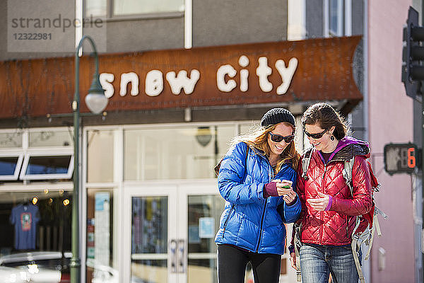 Zwei junge Frauen schauen auf ein Smartphone  während sie vor einem beliebten Restaurant in der Innenstadt von Anchorage  Süd-Zentral-Alaska; Anchorage  Alaska  Vereinigte Staaten von Amerika