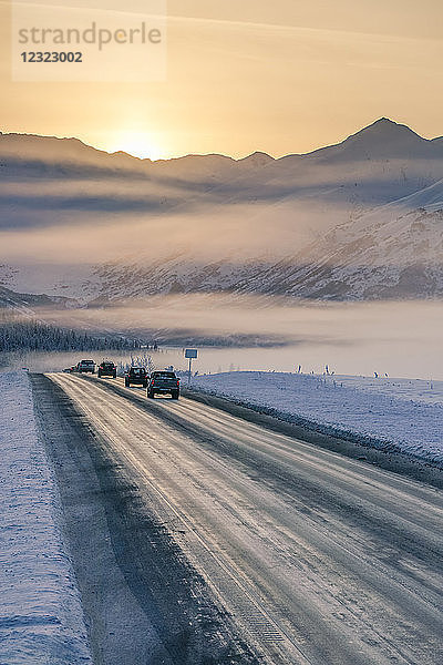 Ein nebliger Sonnenuntergang beleuchtet Autos  die den schneebedeckten Seward Highway hinunterfahren  die Kenai Mountains im Hintergrund  Kenai-Halbinsel  Süd-Zentral-Alaska im Winter; Alaska  Vereinigte Staaten von Amerika