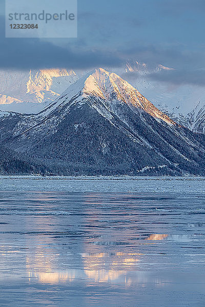 Das warme Licht des Sonnenuntergangs beleuchtet die schneebedeckten Gipfel des Turnagain Passes  die Berge spiegeln sich auf der eisigen Meeresoberfläche  Seward Highway  Süd-Zentral-Alaska im Winter; Alaska  Vereinigte Staaten von Amerika