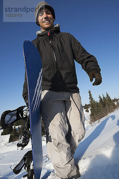 Snowboarder  der mit seiner Ausrüstung im Schnee spazieren geht und lächelnd in die Kamera schaut  Süd-Zentral-Alaska; Homer  Alaska  Vereinigte Staaten von Amerika
