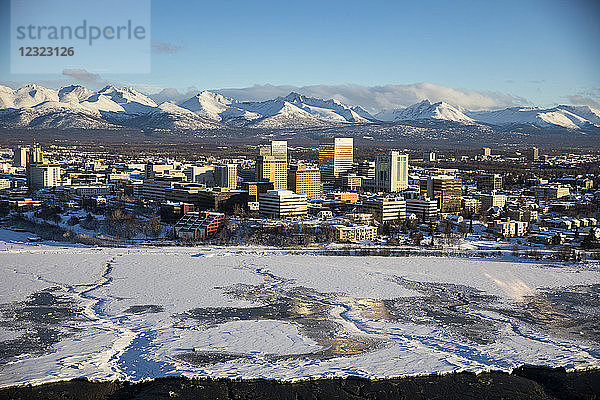 Luftaufnahme von Schnee  der das Meereis an den gefrorenen Ufern der Innenstadt von Anchorage bedeckt  die Chugach Mountains im Hintergrund hinter den Bürogebäuden und Hotels in der Innenstadt von Anchorage im Winter  Süd-Zentral-Alaska; Anchorage  Alaska  Vereinigte Staaten von Amerika