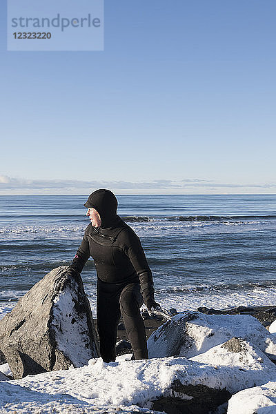 Surferin im Neoprenanzug  die zwischen Felsen und Schnee an der Küste von Homer Spit im Winter spazieren geht  Süd-Zentral-Alaska; Homer Spit  Alaska  Vereinigte Staaten von Amerika