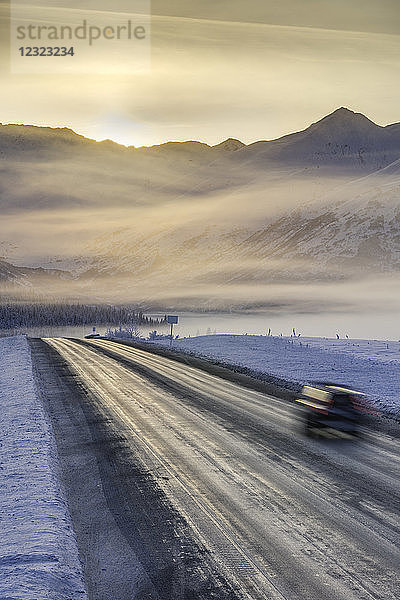 Ein Auto fährt eine Straße in einer schneebedeckten Landschaft hinunter  die Sonne geht über einem Gebirgskamm im Hintergrund auf  Nebel verdunkelt die Silhouette im Winter  Turnagain Pass  Kenai-Halbinsel  Süd-Zentral-Alaska; Alaska  Vereinigte Staaten von Amerika