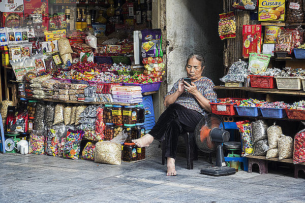 Verkäuferin sitzt barfuß vor ihrem Laden und benutzt ihr Smartphone; Hanoi  Hanoi  Vietnam