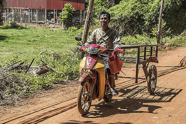 Ein Mann fährt auf einem Motorrad und zieht einen Karren über eine unbefestigte Straße; Siem Reap  Kambodscha