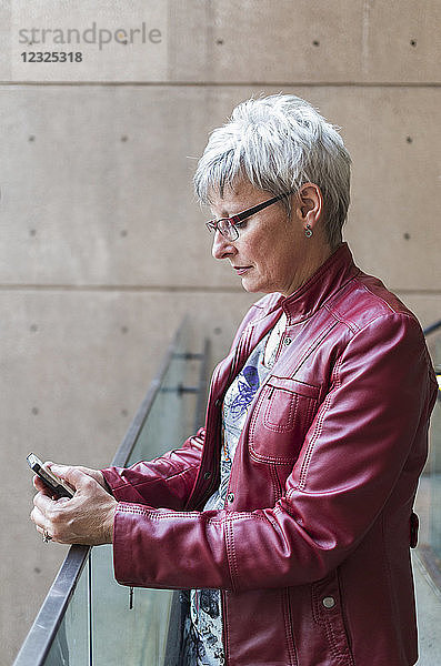 Eine reife Frau in einer roten Lederjacke steht an einem Geländer und benutzt ein Smartphone; Vancouver  British Columbia  Kanada