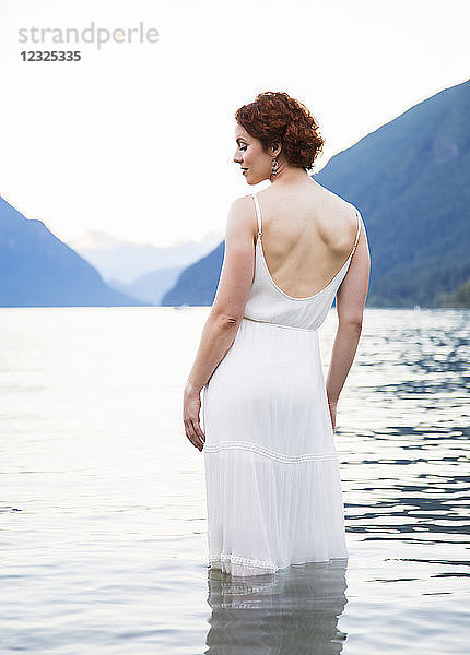 Eine Frau mit roten Haaren und einem weißen Kleid steht an einem Sommerabend im ruhigen Wasser des Alouette Lake; British Columbia  Kanada