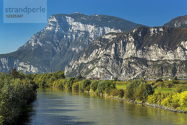Ruhiger Fluss  gesäumt von Bäumen mit grünem Laub und zerklüfteten Bergen im Hintergrund mit blauem Himmel; Trento  Trento  Italien