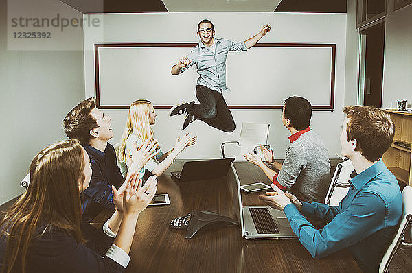 Ein junger Geschäftsmann springt  um eine Leistung mit seinen Mitarbeitern zu feiern  die während einer Präsentation in einem Konferenzraum arbeiten  während einer seiner Mitarbeiter ein Foto mit einem Pad macht; Sherwood Park  Alberta  Kanada