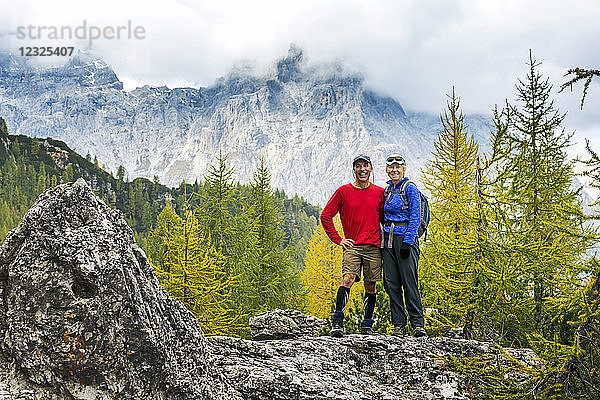 Wanderer und Wanderin stehen auf einem Felsen mit herbstlich gefärbten Bäumen und einer wolkenbedeckten Bergkette im Hintergrund; Sexten  Bozen  Italien