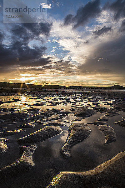 Nasse Wellen im Sand an einem Strand bei Sonnenuntergang mit einem Sonnenaufgang über silhouettierten Hügeln in der Ferne; Embleton  Northumberland  England
