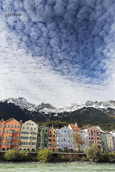 Bunte Gebäude am Flussufer mit schneebedeckten Berggipfeln  dramatischen Wolken und blauem Himmel über dem Wasser; Innsbruck  Tirol  Österreich