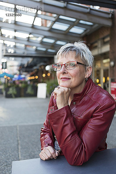 Porträt einer reifen Frau in einer roten Lederjacke  die an einem Tisch vor einem Geschäft sitzt und ihr Kinn in die Hand stützt; Vancouver  British Columbia  Kanada