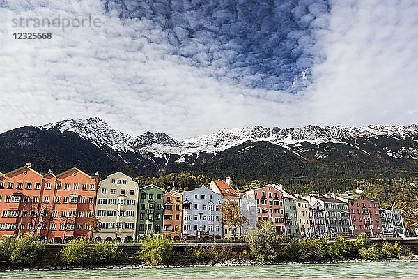 Bunte Gebäude entlang des Flussufers mit schneebedeckten Berggipfeln  dramatischen Wolken und blauem Himmel über dem Wasser; Innsbruck  Tirol  Österreich