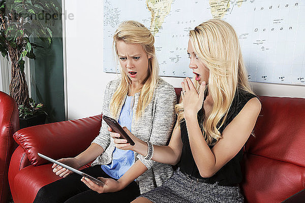 Zwei hübsche junge Geschäftsfrauen der Millennials sitzen an einem Arbeitsplatz und nutzen ihre Technologie mit schockierten Gesichtern  während sie etwas in den sozialen Medien lesen; Sherwood Park  Alberta  Kanada