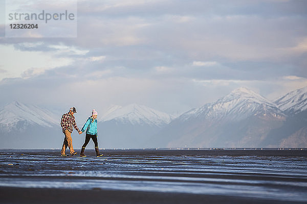 Ein junges Paar  das an einem Strand spazieren geht und sich an den Händen hält  mit einer Bergkette in der Ferne; Anchorage  Alaska  Vereinigte Staaten von Amerika