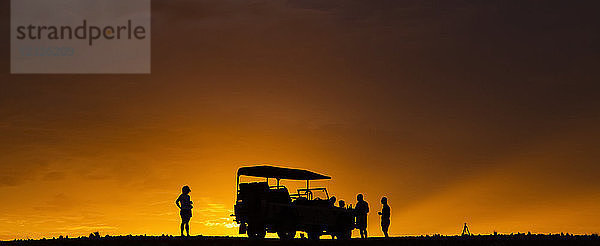 Silhouette von Touristen auf einer Luxus-Safari  die neben einem Freizeitfahrzeug in der Wüste bei Sonnenuntergang stehen; Sossusvlei  Hardap-Region  Namibia