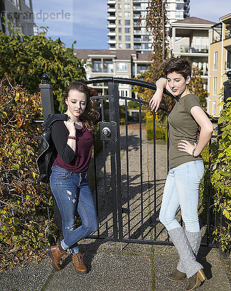 Zwei junge Frauen stehen an ein Metalltor gelehnt und sehen selbstbewusst und cool aus  mit einem Haus im Hintergrund; New Westminster  British Columbia  Kanada