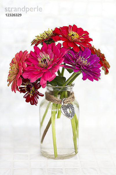 Eine Vielzahl farbiger Zinnienblüten in einer schlichten Glasvase mit einem dekorativen Herzanhänger  der mit einer Juteschnur befestigt ist