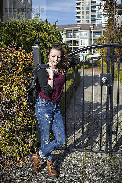 Eine junge Frau steht an ein Metalltor gelehnt  hält eine Lederjacke in der Hand und sieht zuversichtlich aus  mit einem Haus im Hintergrund; New Westminster  British Columbia  Kanada